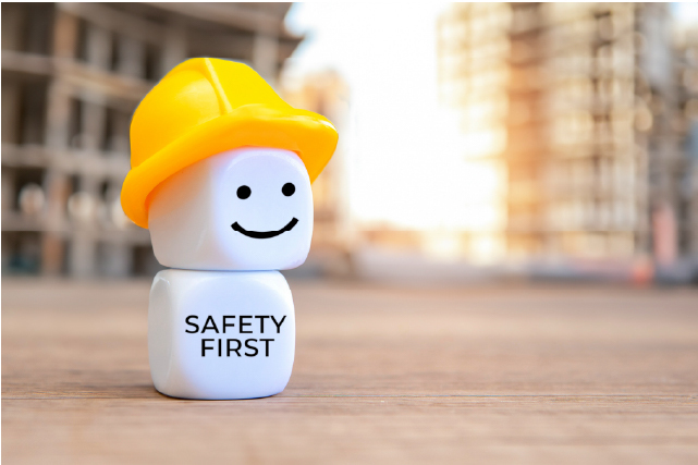 Cute construction safety figure with yellow helmet and “Safety First” text on a building site background.